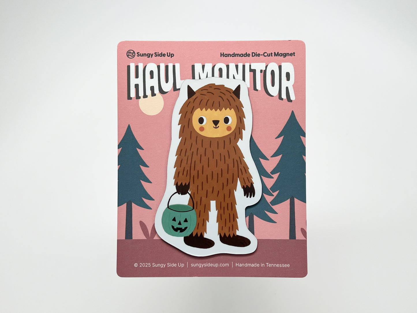 Handmade die-cut Bigfoot Halloween magnet on illustrated backing card with a pink forest background. Features a cute brown sasquatch holding a teal jack-o’-lantern candy bucket. Perfect quirky fridge décor for cryptid fans, spooky season gifts, and woodland creature collectors.