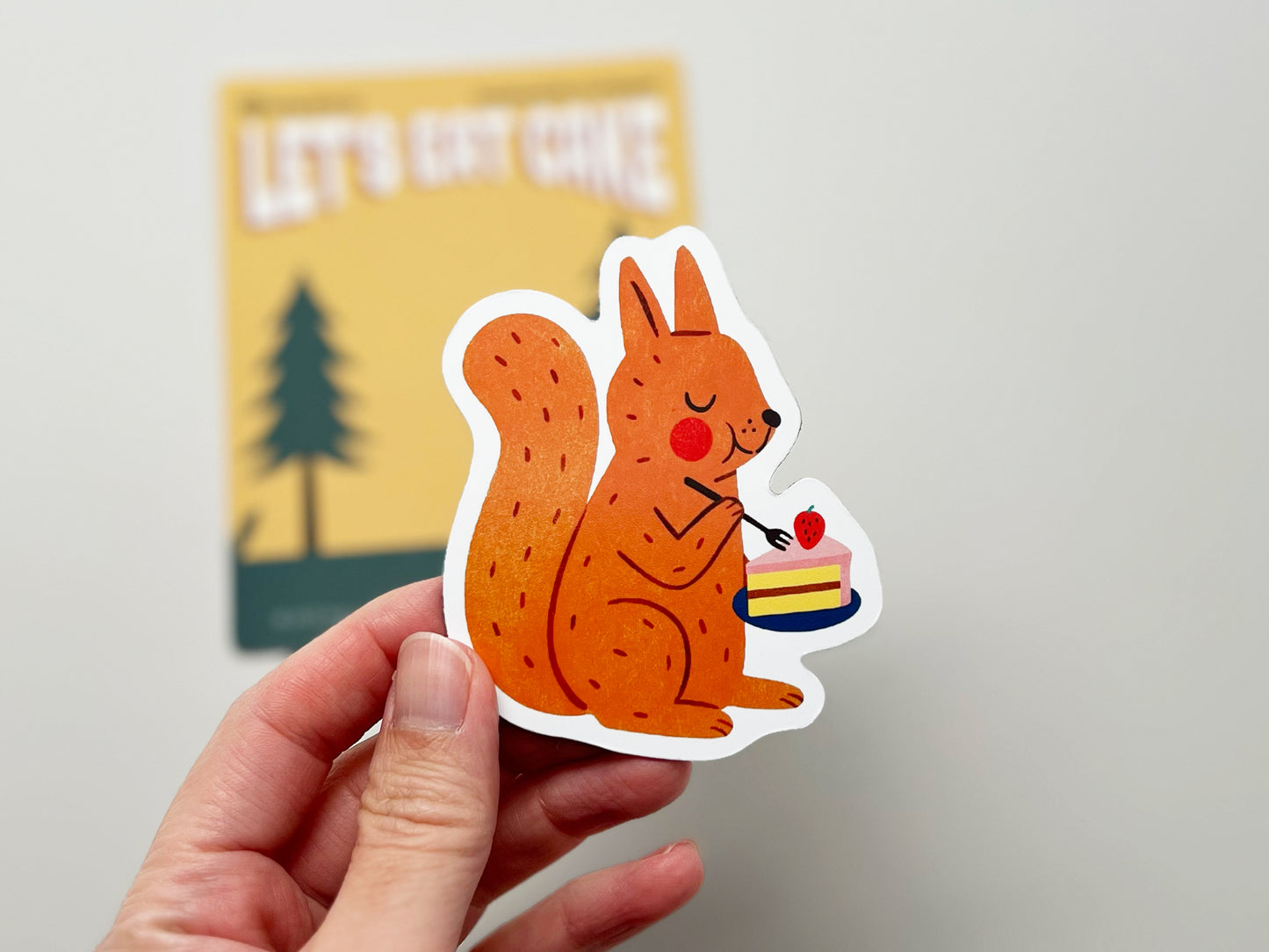 Close-up of a handmade die-cut fridge magnet featuring a cute illustrated red squirrel holding a plate with a slice of strawberry-topped cake. The vibrant woodland animal design is perfect for nature lovers, dessert fans, and quirky kitchen or cabin decor.