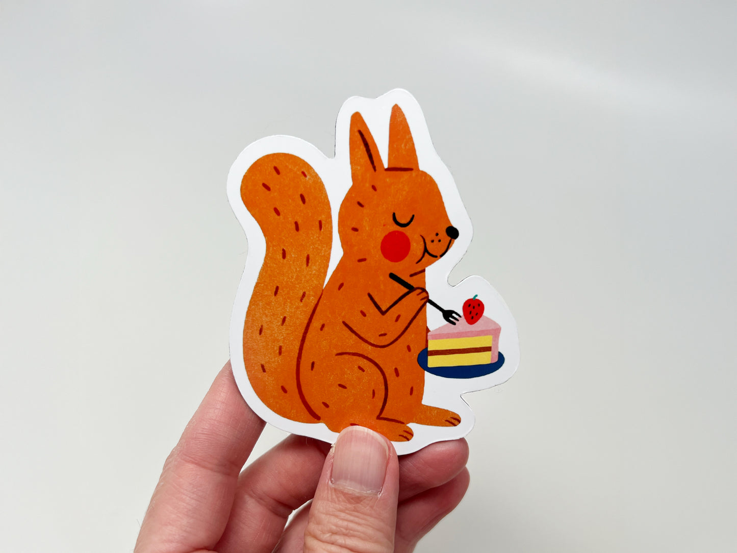 Hand holding a handmade die-cut fridge magnet shaped like an orange squirrel enjoying a slice of strawberry-topped cake. The cute woodland animal design is vibrant and playful, making it ideal for nature lovers, dessert fans, and quirky kitchen or home decor.