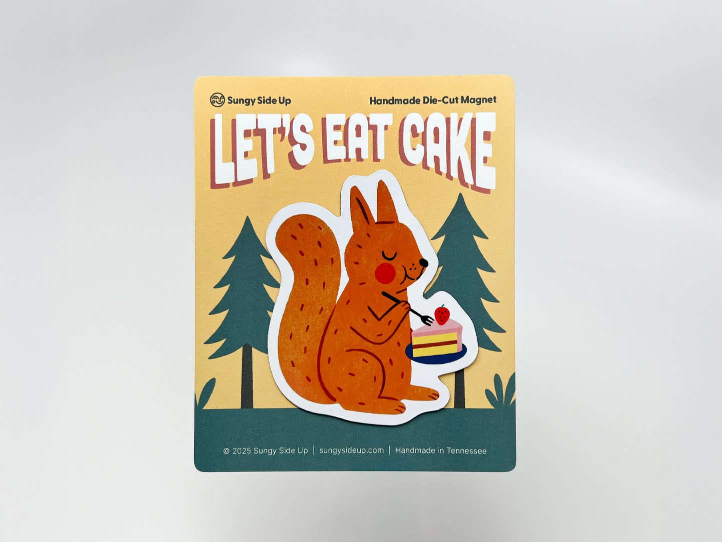 Handmade die-cut fridge magnet featuring an illustrated orange squirrel holding a plate with a slice of strawberry-topped cake, mounted on decorative backing with a woodland tree design. Perfect for animal lovers, dessert fans, and quirky kitchen or cabin decor.