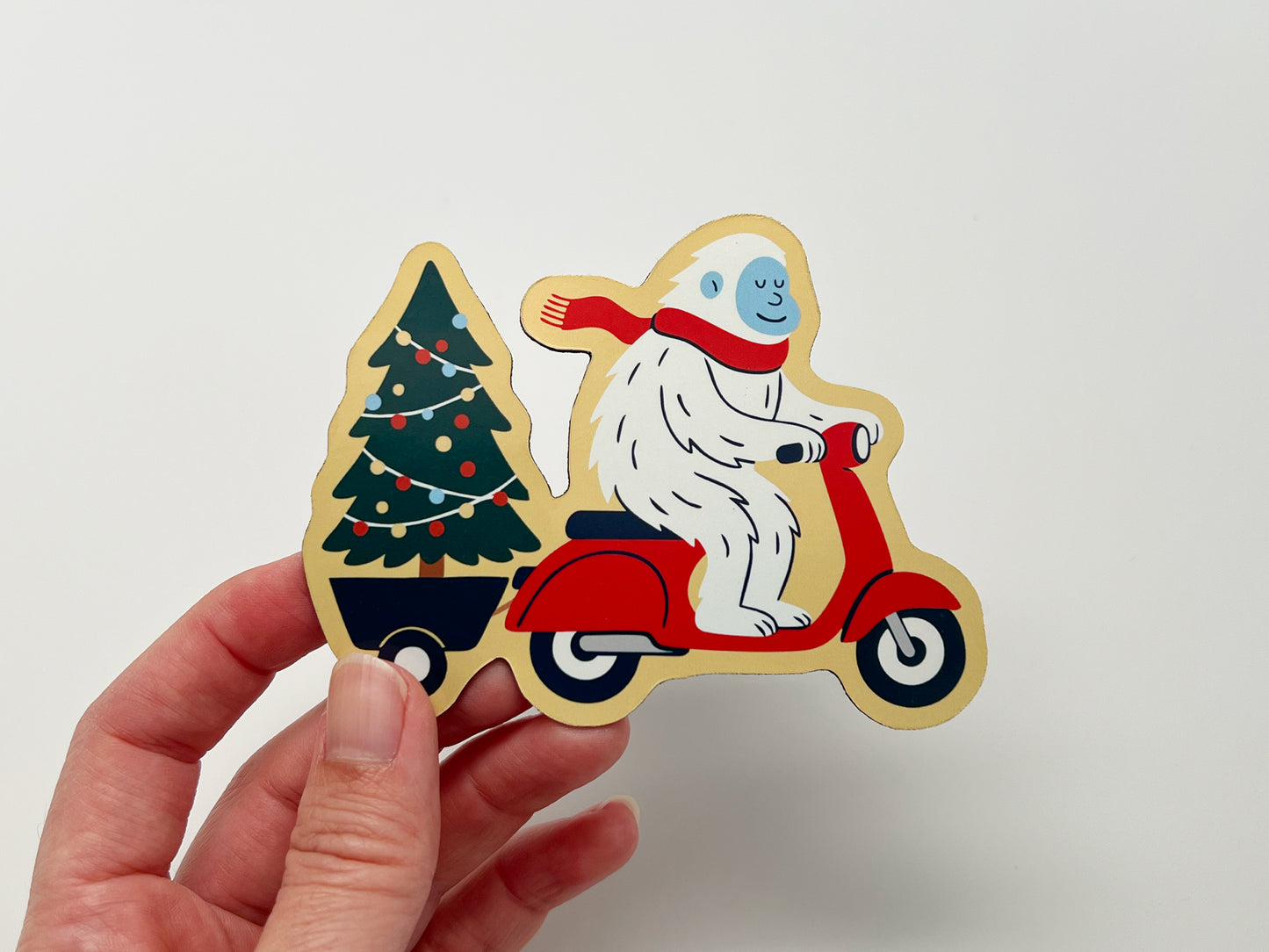 Hand holding a handmade die-cut holiday magnet shaped like a smiling yeti in a red scarf riding a red scooter while towing a decorated Christmas tree. The quirky magnet has bold colors and clean lines, perfect as festive décor, stocking stuffer, or unique holiday gift for fridge or office.