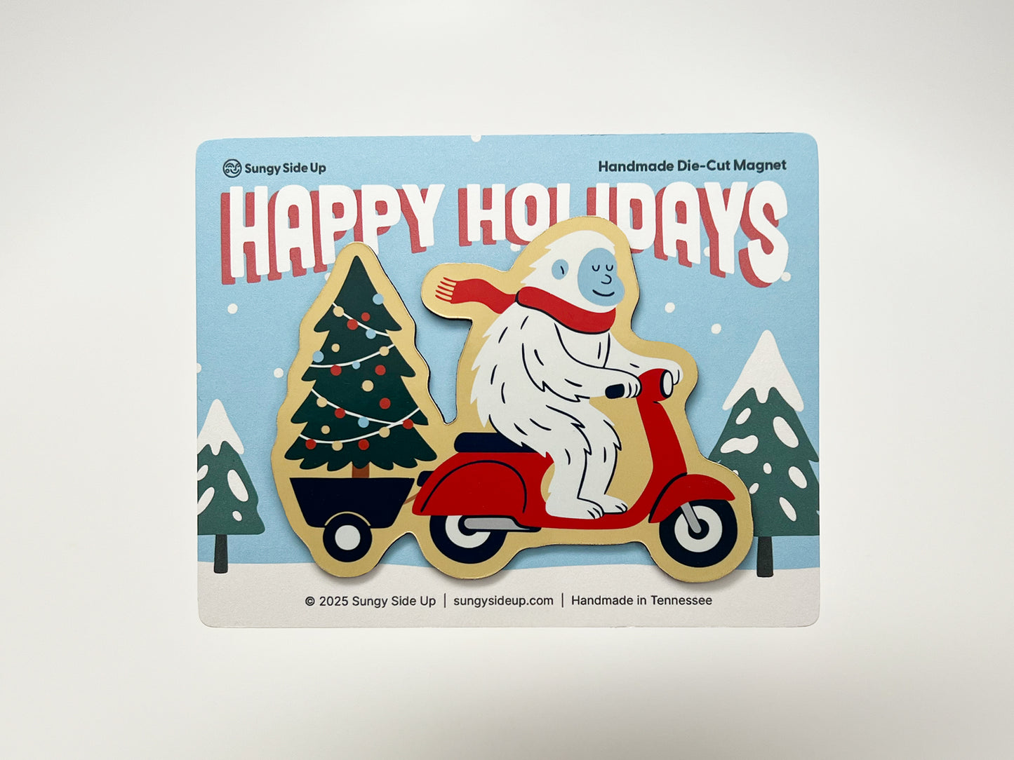 Handmade die-cut holiday magnet featuring a cheerful yeti riding a bright red scooter, hauling a decorated Christmas tree on a snowy background. The festive backing card says “Happy Holidays” in bold red letters. A quirky and fun Christmas decoration, perfect for gifting or holiday décor.