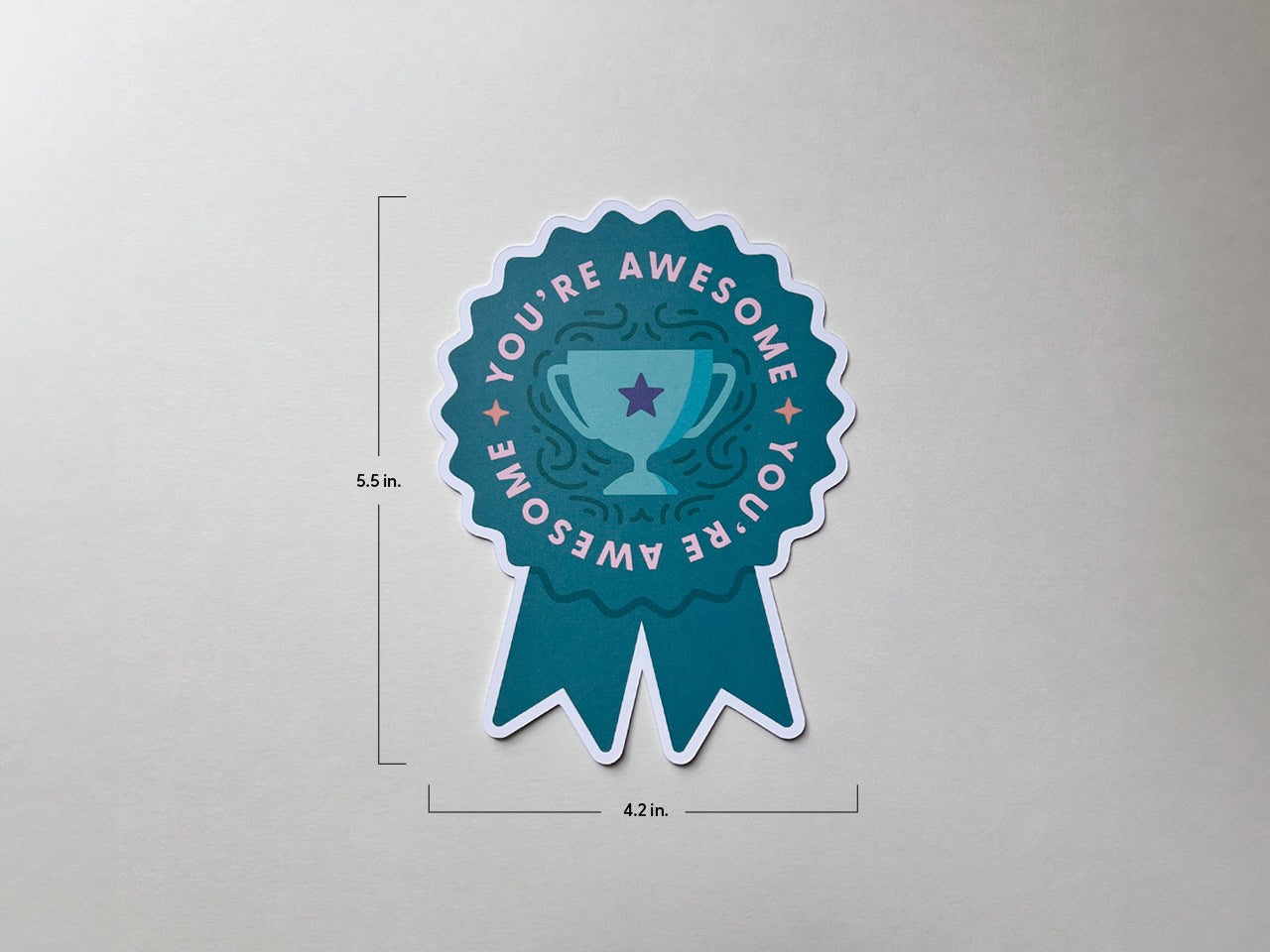 A teal die-cut note card shaped like a prize ribbon is displayed on a neutral background. It measures 5.5 inches tall and 4.2 inches wide, with You're Awesome written around a trophy illustration.