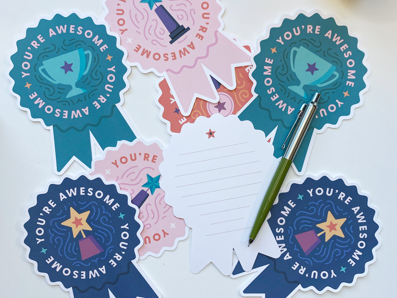 A collection of die-cut note cards shaped like prize ribbons is laid out on a white surface. Each card displays the phrase You're Awesome with illustrations of trophies. One card is flipped to its back, lined for writing a personal message.