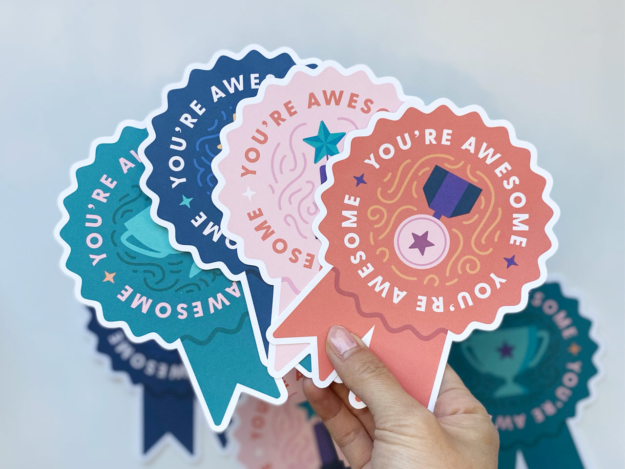 A hand holds a set of colorful die-cut note cards shaped like prize ribbons. Each card features the phrase You're Awesome and a trophy illustration. The cards are in colors indigo blue, teal green, and pink. The background is a light, neutral tone.