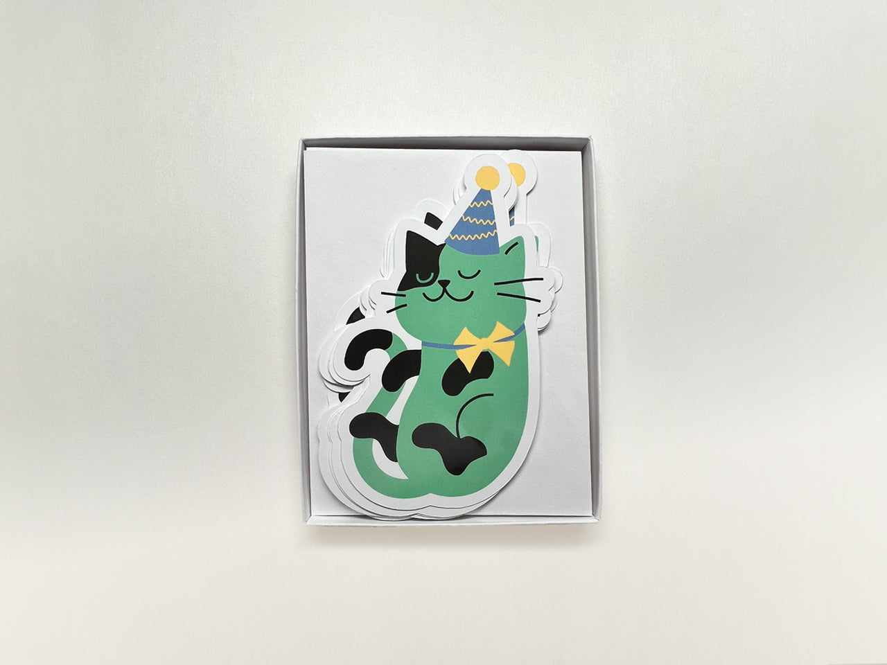 A box of cat-shaped note cards featuring a green cat illustration wearing a purple party hat and yellow bowtie. The set of note cards are stacked neatly with white envelopes underneath, perfect for gifting and celebrations.