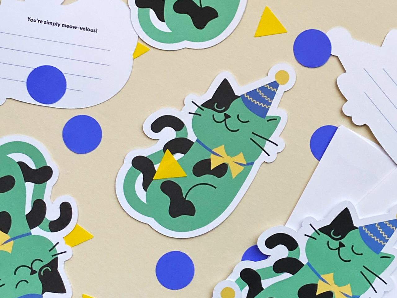 A flat-lay arrangement of multiple party-cat note cards scattered across a table. The note cards feature a cheerful green cat with a blue and yellow party hat and a bright yellow bowtie.