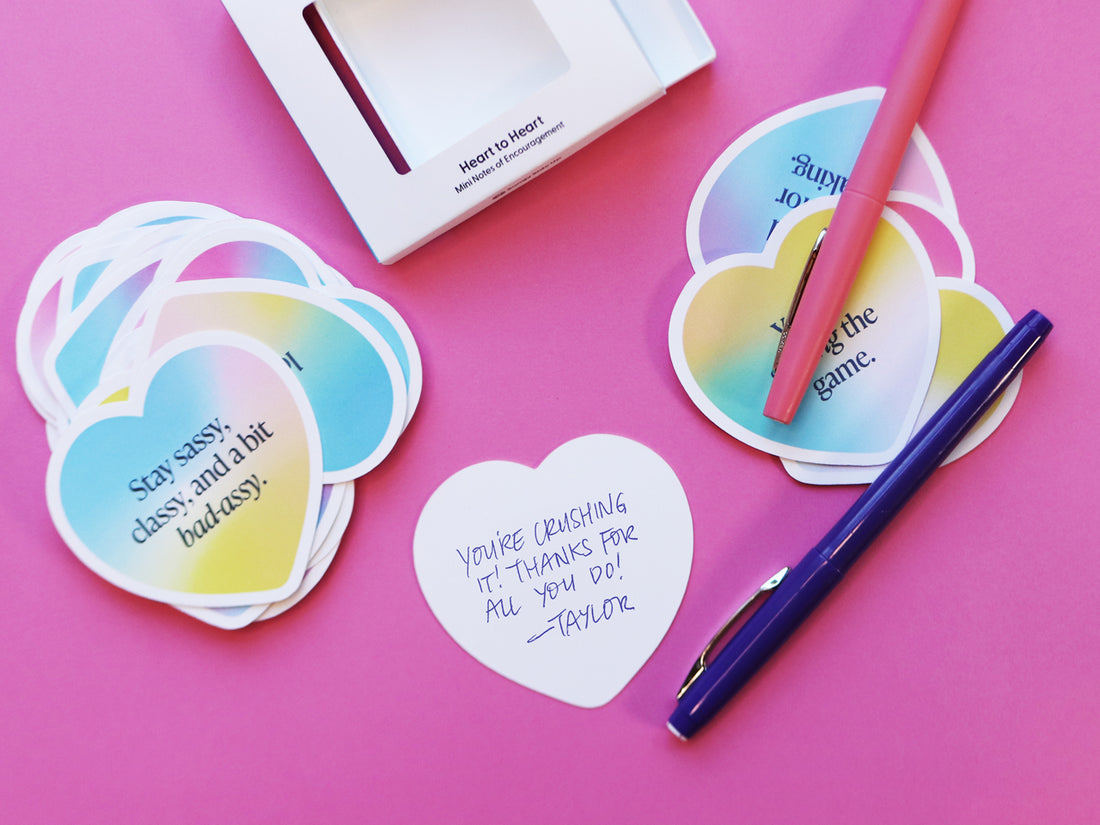 Colorful heart-shaped encouragement notes on a pink background, featuring positive messages like "Stay sassy, classy, and a bit bad-assy." A handwritten note reads, "You're crushing it! Thanks for all you do! - Taylor," with pink and blue pens nearby.