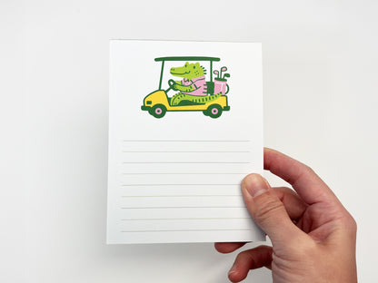 Hand holding a cute alligator golf cart notepad, 4.25 x 5.5 inch lined memo pad with an illustrated animal design on a white background.