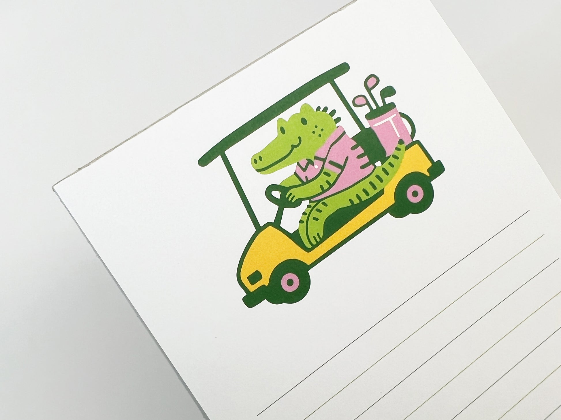 Close-up of a cute illustrated alligator driving a yellow golf cart at the top of a lined 4.25 x 5.5 inch notepad, playful animal stationery design on white paper.