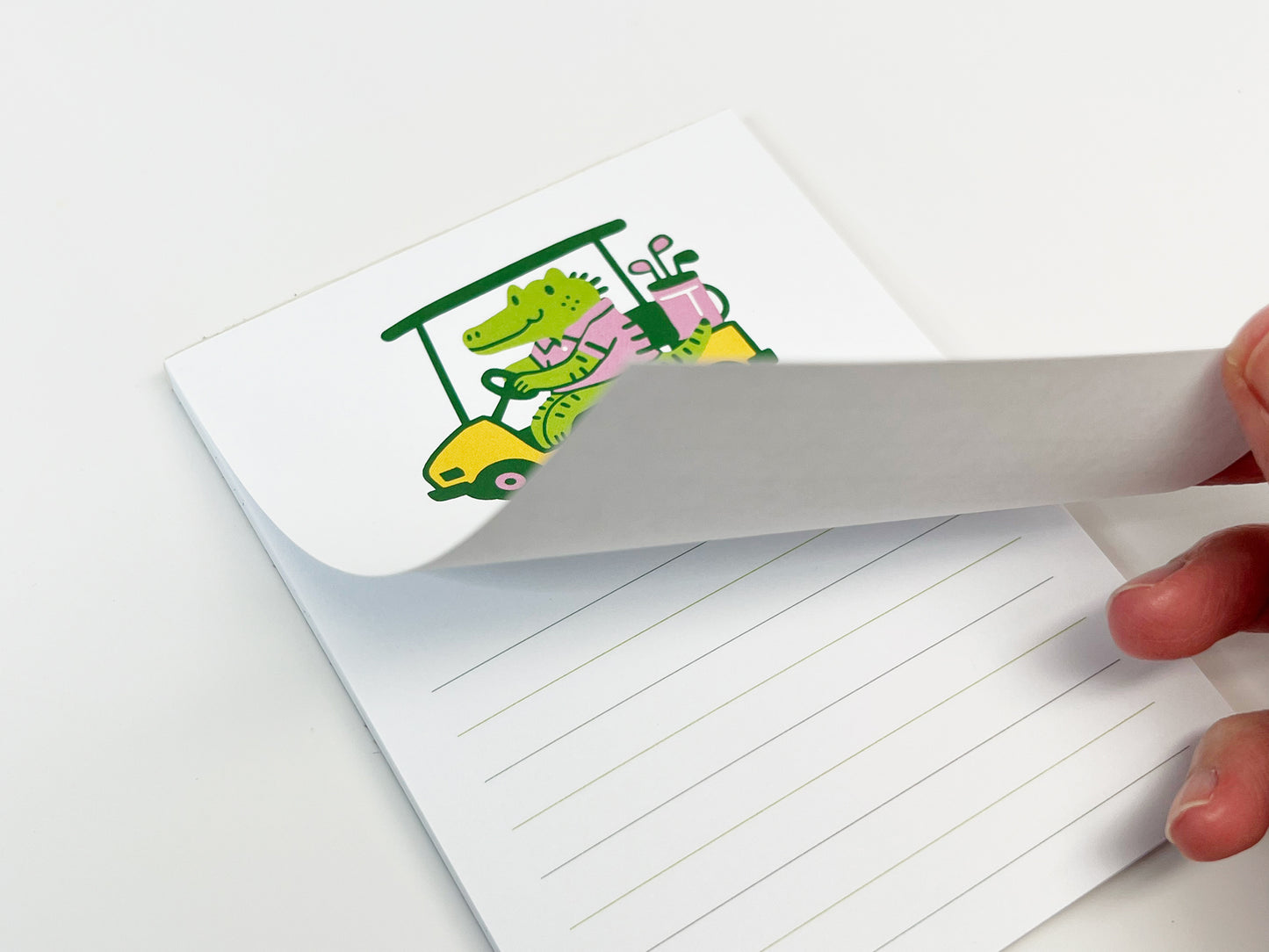 Close-up of a lined alligator golf cart notepad with a page being lifted to show paper thickness, featuring a cute illustrated animal design on a 4.25 x 5.5 inch memo pad.