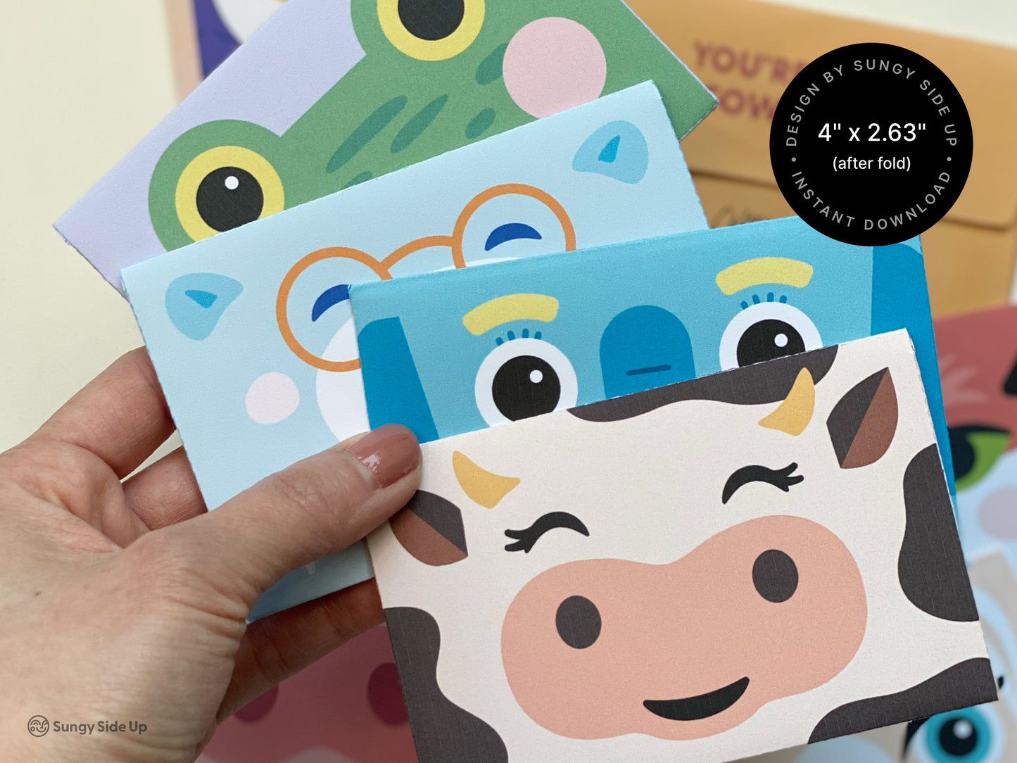 A collection of animal-themed printable gift card holders is shown, featuring designs like a winking cow, a frog with wide yellow eyes, and a blue elephant with playful details. The envelopes are colorful, sized at approximately 4" x 2.63" when folded, and designed for easy assembly at home. A hand holds some of the assembled gift card holders, highlighting their vibrant colors and cute expressions. These printables are ideal for kids’ birthdays, parties, and creative gifting.