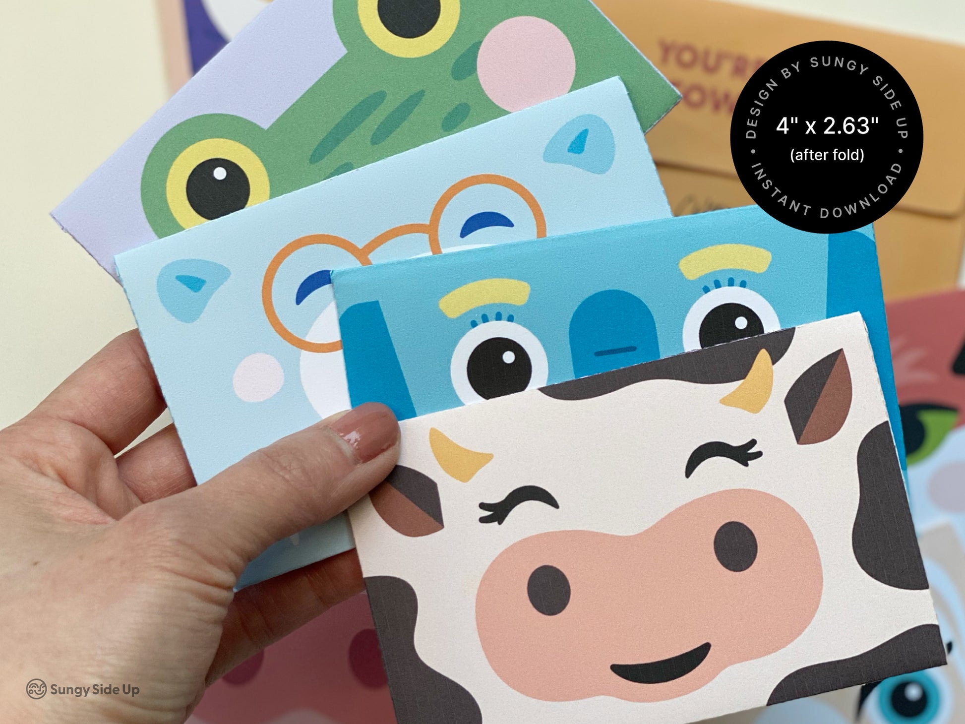 A collection of animal-themed printable gift card holders is shown, featuring designs like a winking cow, a frog with wide yellow eyes, and a blue elephant with playful details. The envelopes are colorful, sized at approximately 4" x 2.63" when folded, and designed for easy assembly at home. A hand holds some of the assembled gift card holders, highlighting their vibrant colors and cute expressions. These printables are ideal for kids’ birthdays, parties, and creative gifting.