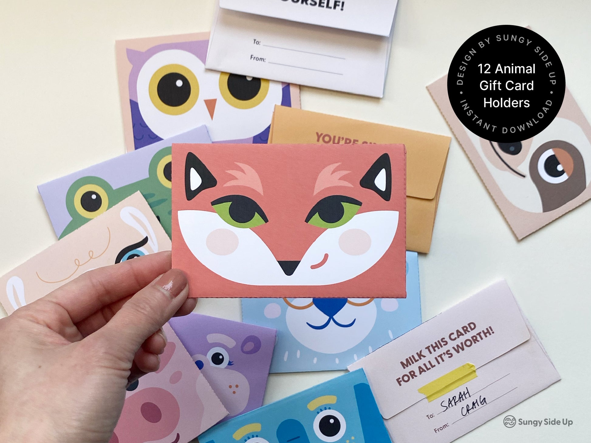 A hand holds a colorful printable animal-themed gift card holder featuring a bright fox design with green eyes and a white face. Other animal designs, including a frog, owl, and sloth, are scattered on the surface, each cut, folded, and ready to use. The printable cards are designed for DIY assembly at home, formatted for US Letter size, and perfect for creating fun, personalized envelopes for birthdays, parties, and special occasions. A playful saying appears on the back of each card.
