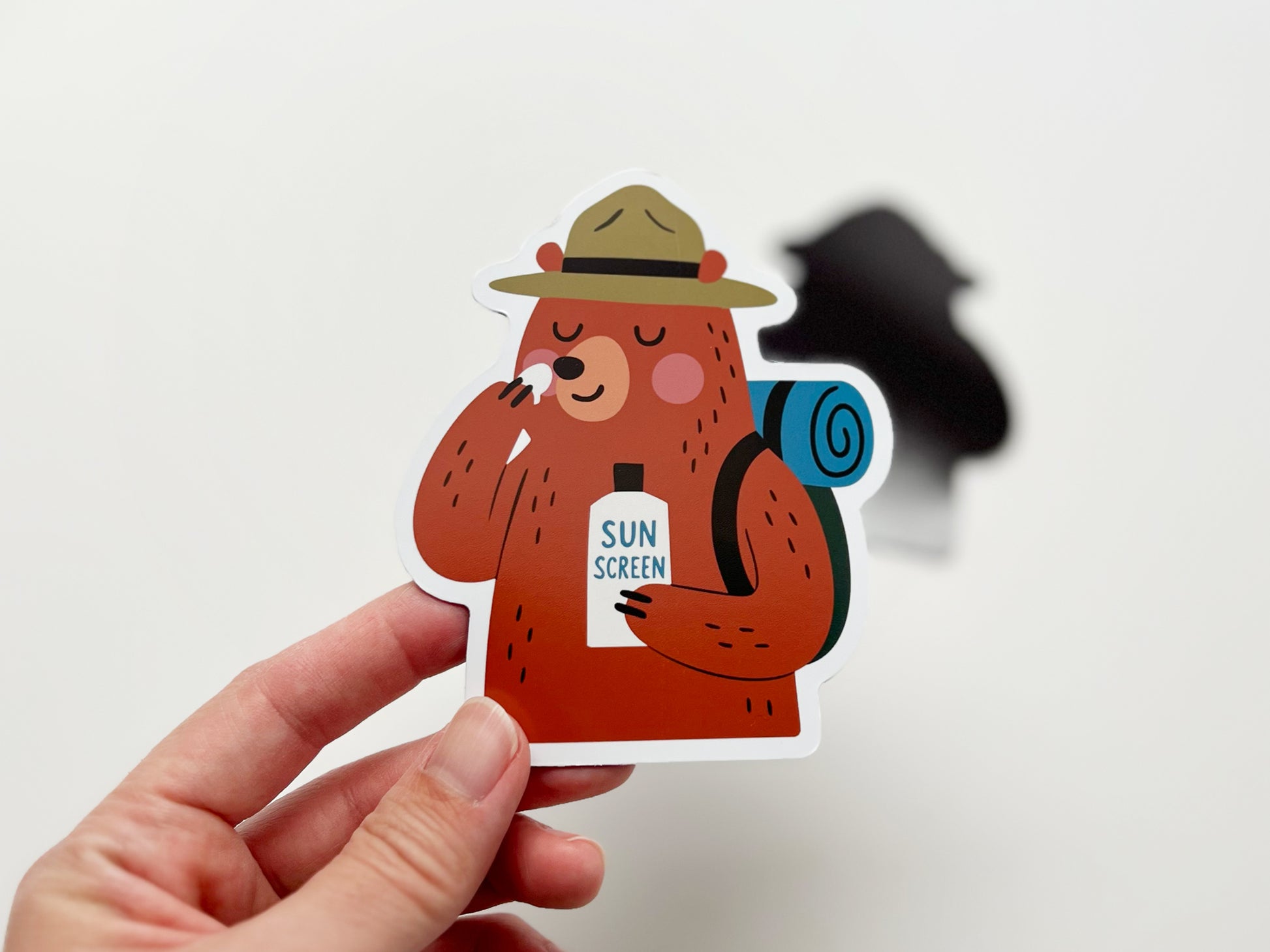 Hand holding a handmade die-cut fridge magnet shaped like a cartoon bear camper. The brown bear wears a ranger hat, carries a blue rolled backpack, and applies sunscreen to its nose while holding a bottle labeled “SUN SCREEN.” Quirky camping magnet gift for hikers, campers, and outdoor lovers.
