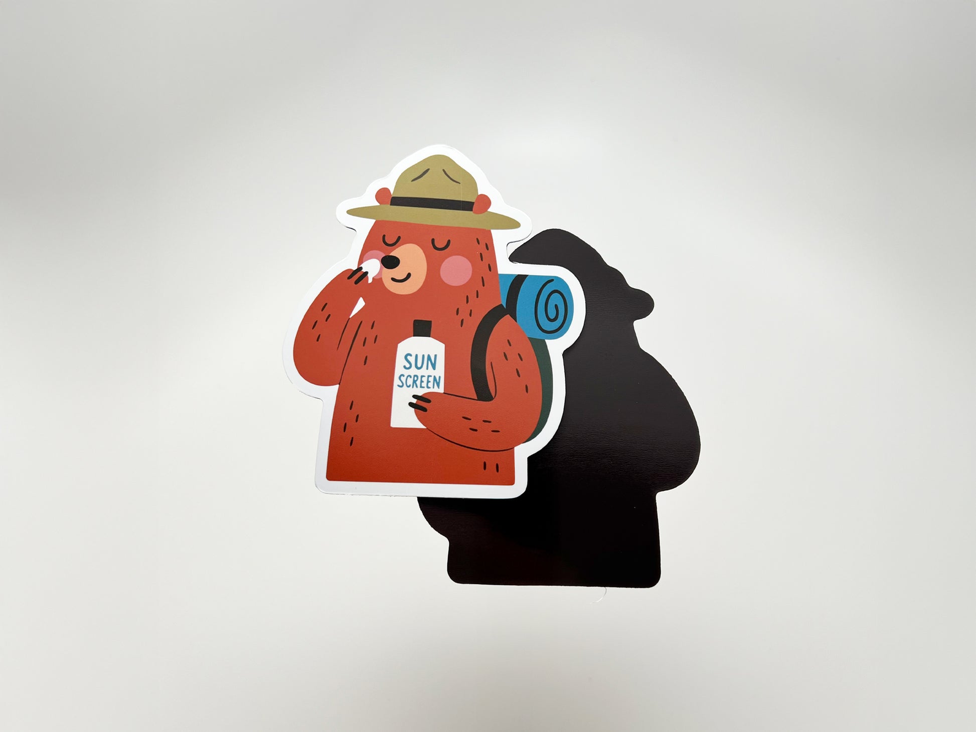 Handmade die-cut magnet featuring a cartoon bear camper with a ranger hat and blue backpack, smiling while applying sunscreen from a bottle labeled “SUN SCREEN.” The magnet is displayed on a plain background with its black silhouette shadow visible. Fun camping gift for hikers and outdoor lovers.
