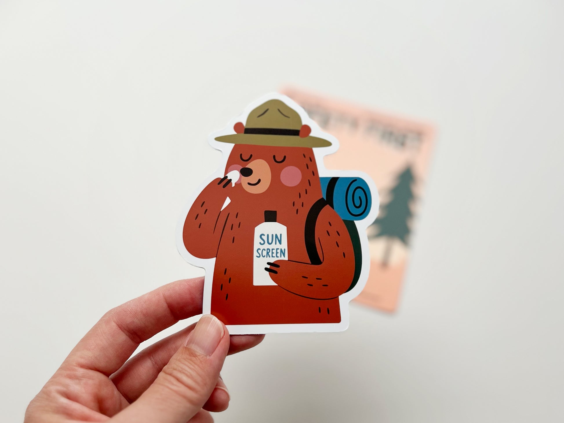 Hand holding a handmade die-cut fridge magnet of a cartoon bear camper. The bear wears a ranger hat, carries a blue rolled backpack, and applies sunscreen to its face with a bottle labeled “SUN SCREEN.” A peach “Safety First” backing card with trees is blurred in the background. Fun camping gift idea.