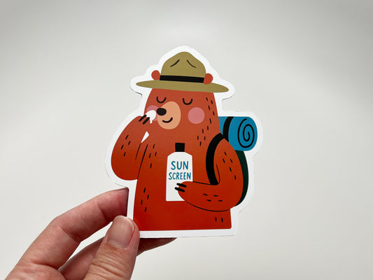 Close-up of a handmade die-cut fridge magnet featuring a brown bear camper. The bear wears a ranger hat, carries a rolled blue backpack, and applies sunscreen with a bottle labeled “SUN SCREEN.” Fun outdoorsy bear magnet gift for hikers, campers, and quirky cabin or kitchen decor.