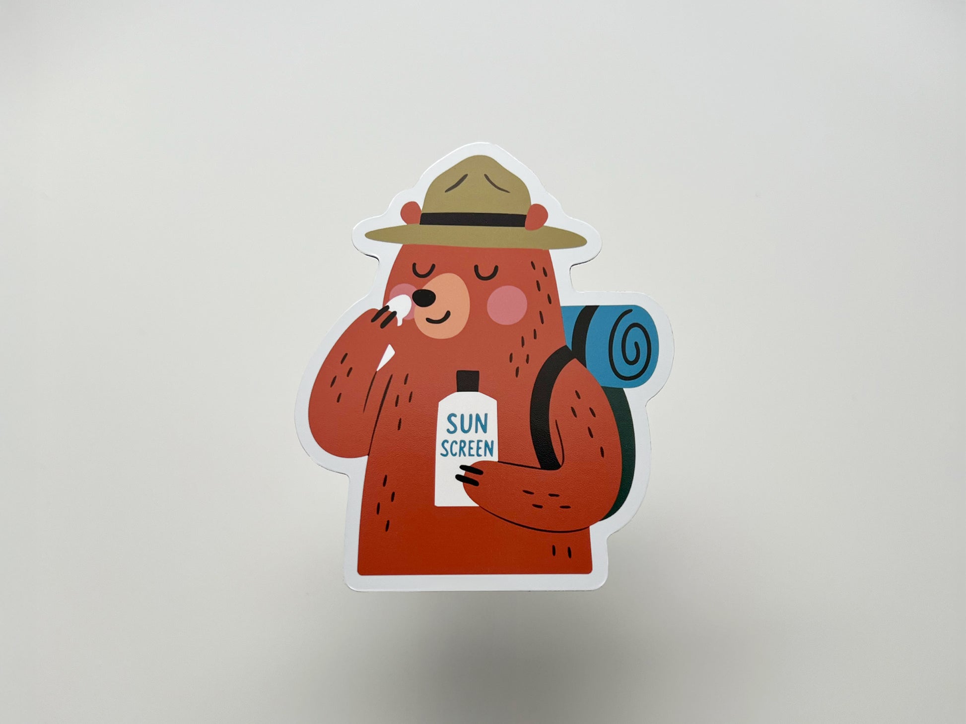 Handmade die-cut fridge magnet featuring a cartoon bear camper with a ranger hat and blue rolled backpack. The smiling brown bear applies sunscreen from a bottle labeled “SUN SCREEN.” Cute and quirky camping magnet gift for hikers, backpackers, national park lovers, or fun cabin kitchen decor.