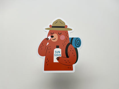 Handmade die-cut fridge magnet featuring a cartoon bear camper with a ranger hat and blue rolled backpack. The smiling brown bear applies sunscreen from a bottle labeled “SUN SCREEN.” Cute and quirky camping magnet gift for hikers, backpackers, national park lovers, or fun cabin kitchen decor.