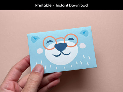 A hand holding a cute printable bear gift card holder featuring a smiling bear face with orange glasses on a light blue background. The bear illustration is cheerful and whimsical, perfect for kids' birthdays, baby showers, or teacher appreciation gifts. This digital download is easy to print and assemble at home, making your gift card presentation extra special and fun for any celebration or occasion.