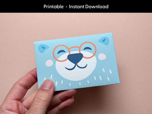 A hand holding a cute printable bear gift card holder featuring a smiling bear face with orange glasses on a light blue background. The bear illustration is cheerful and whimsical, perfect for kids' birthdays, baby showers, or teacher appreciation gifts. This digital download is easy to print and assemble at home, making your gift card presentation extra special and fun for any celebration or occasion.