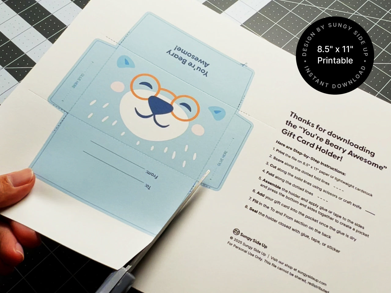 A close-up of a person cutting out the printable bear gift card holder with scissors on a cutting mat. The blue bear design includes fold lines, glue tabs, and a message area. A printed instruction sheet is visible beside the design, guiding users through the simple DIY assembly process. This image shows how easy and hands-on it is to create a fun gift card envelope at home with this instant download—perfect for last-minute gift giving.