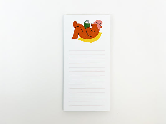 Vertical lined notepad on a white background featuring an illustration at the top of a brown bear lounging on a yellow cushion while reading a green book, wearing a pink hat; light pink horizontal lines fill the lower portion of the notepad for writing.