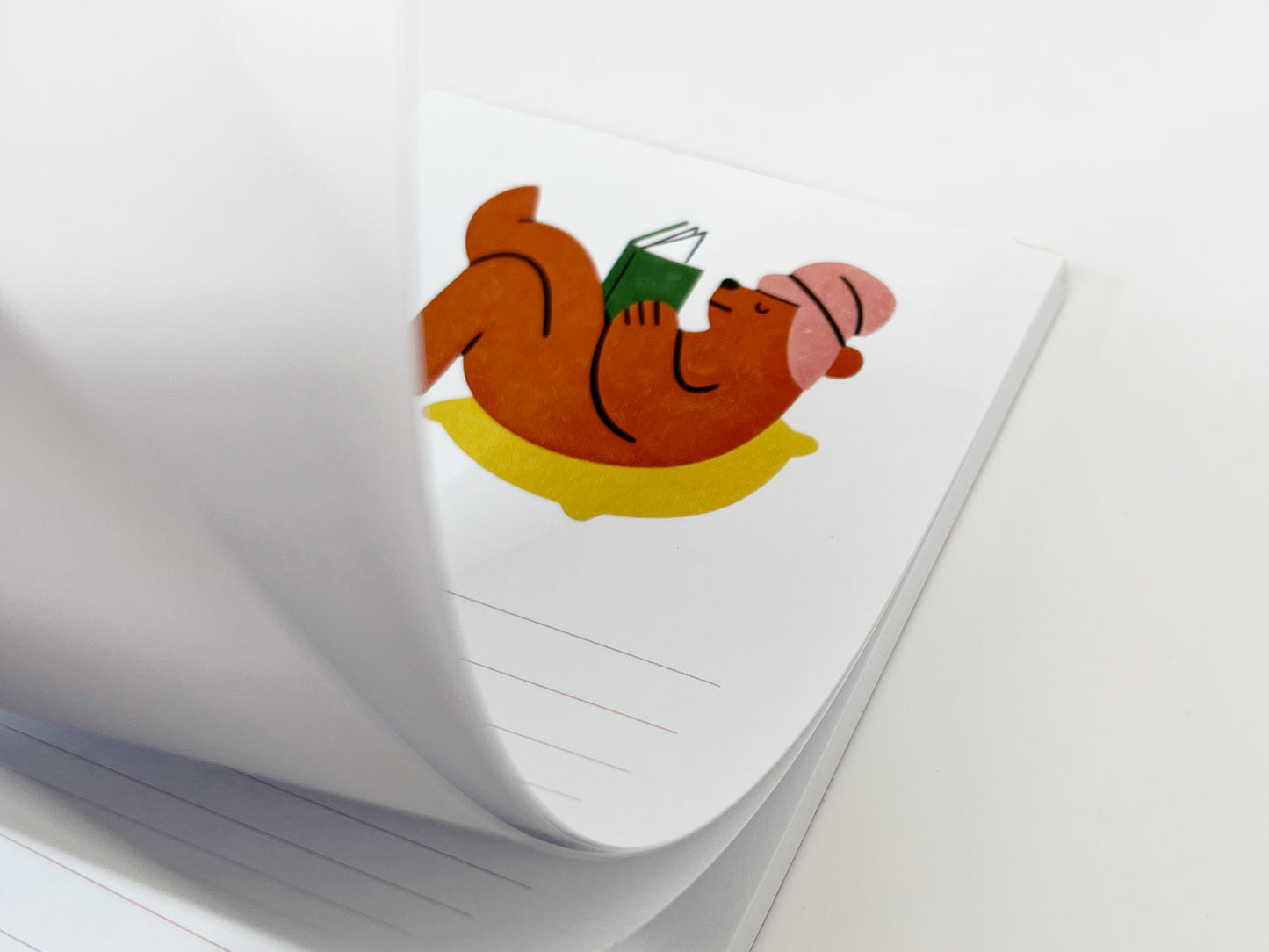Close-up angled view of a lined notepad with pages partially flipped, showing an illustration of a brown bear lounging on a yellow cushion while reading a green book and wearing a pink hat; light pink horizontal lines are visible on the pages against a white background.