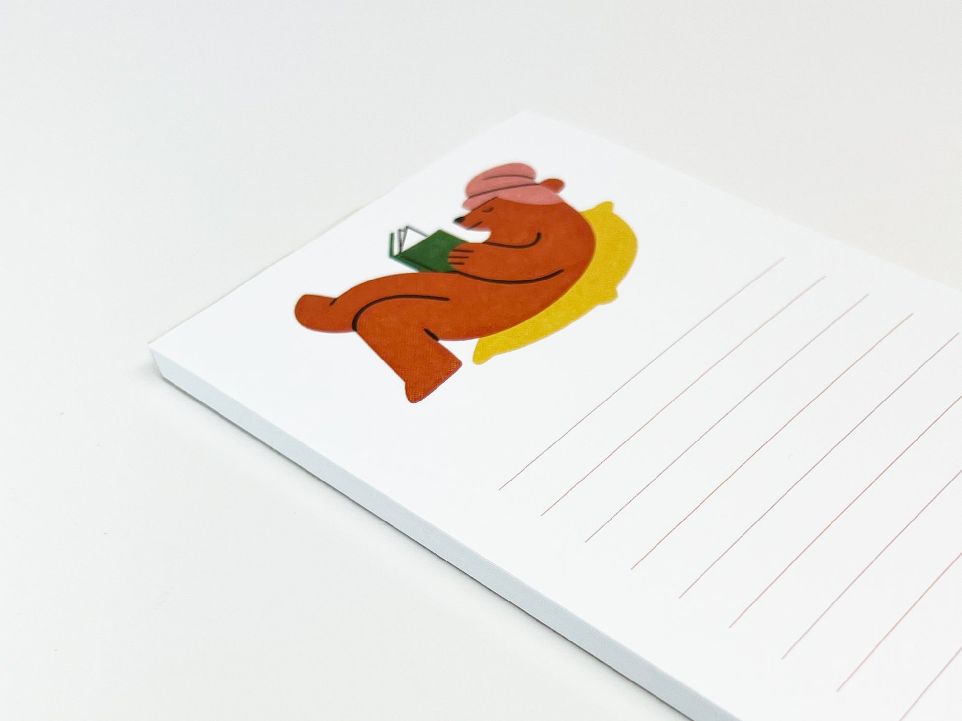 Angled close-up of a slim lined notepad on a white surface, featuring an illustration of a brown bear reclining on a yellow cushion while reading a green book and wearing a pink hat; light pink horizontal lines appear on the lower portion of the page.
