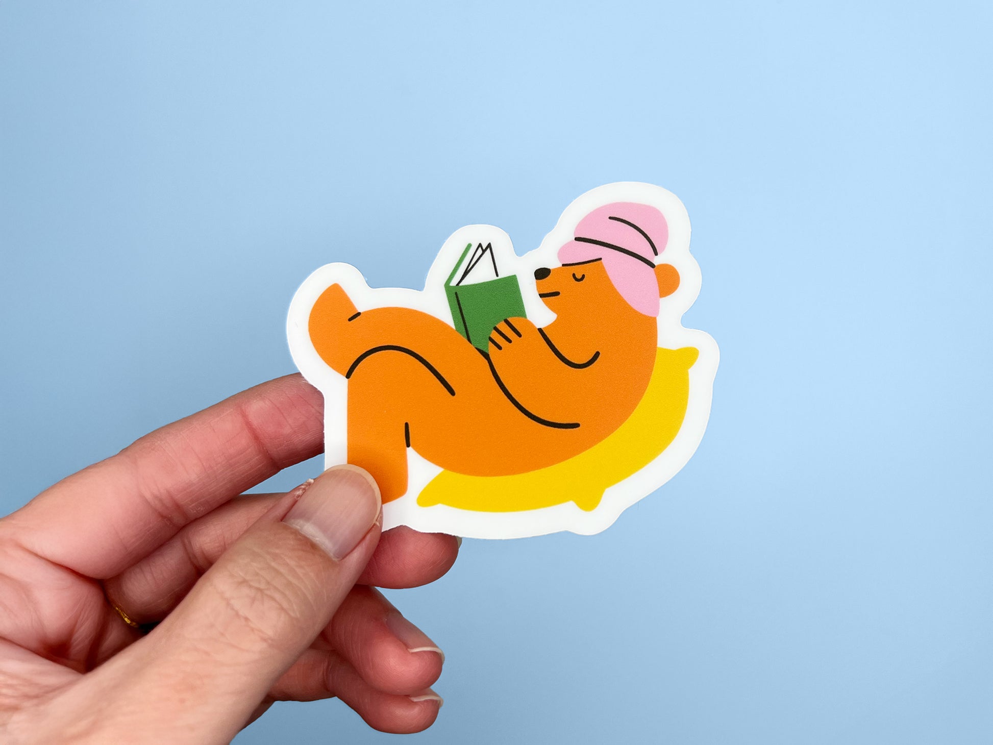 Hand holding a matte vinyl die cut sticker of an orange bear wearing a pink towel around its head, lying on a yellow pillow while reading a green book. The cozy reading bear sticker is shown against a light blue background.