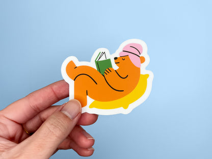 Hand holding a matte vinyl die cut sticker of an orange bear wearing a pink towel around its head, lying on a yellow pillow while reading a green book. The cozy reading bear sticker is shown against a light blue background.