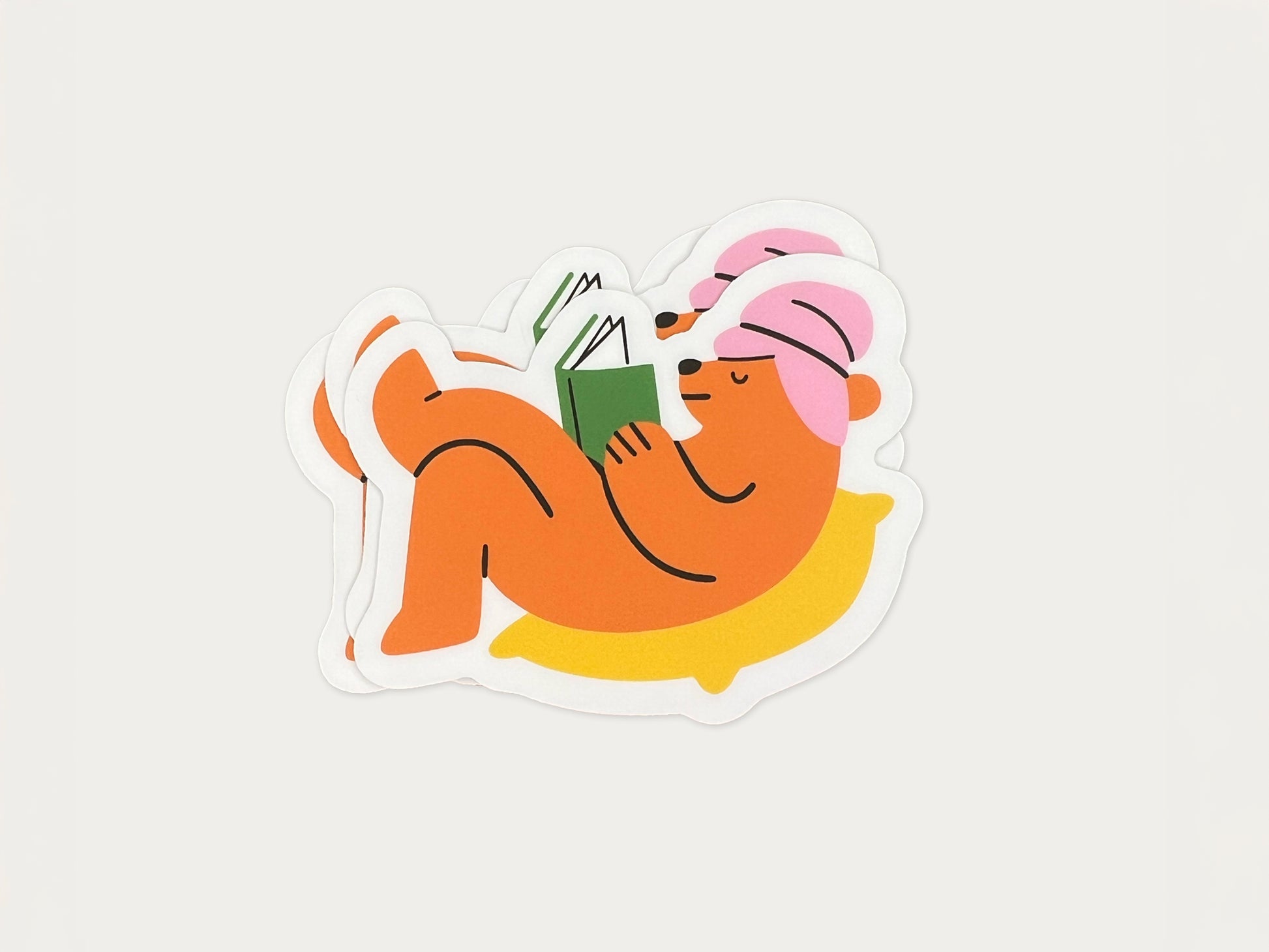 Small stack of matte vinyl die cut stickers showing an orange bear wearing a pink towel around its head, lying on a yellow pillow while reading a green book. Multiple cozy reading bear stickers are layered together on a light neutral background.