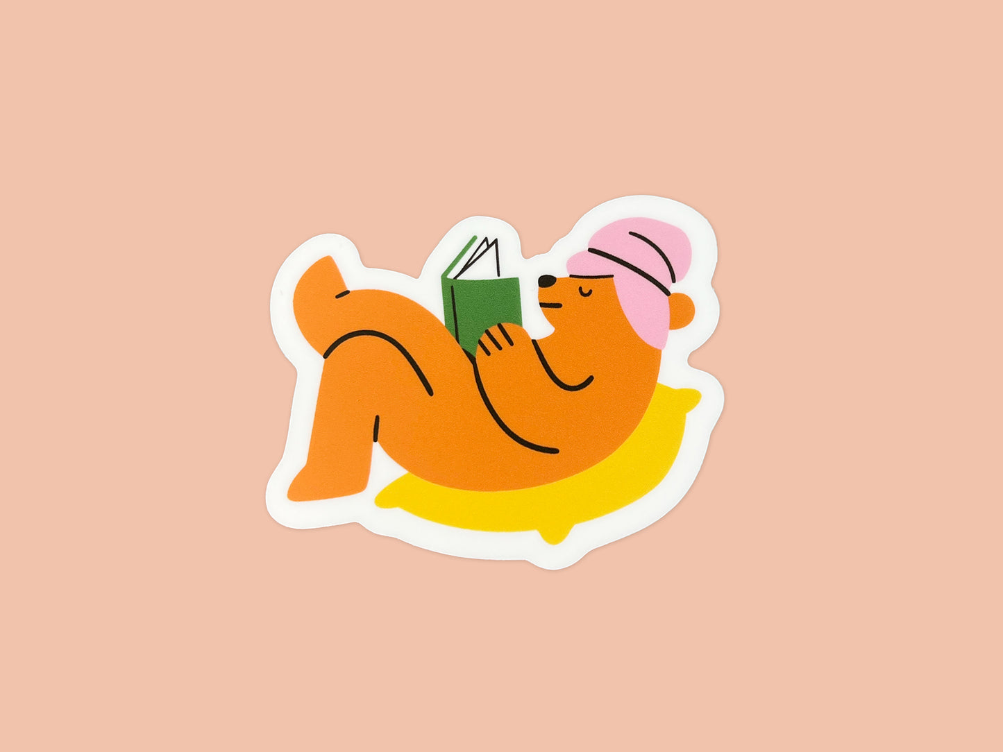 Single matte vinyl die cut sticker of an orange bear wearing a pink towel wrapped around its head, lying on a yellow pillow while holding and reading a green book. The cozy reading bear sticker is centered on a soft peach background.