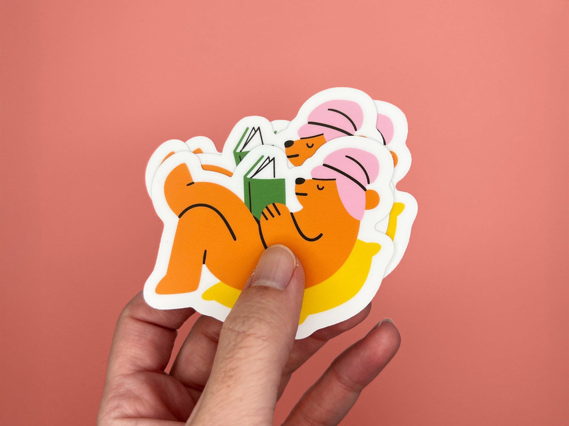 Hand holding a small stack of matte vinyl die cut stickers showing an orange bear wearing a pink towel around its head, lying on a yellow pillow while reading a green book. Multiple stickers are layered together against a coral background, highlighting the cozy reading bear design.