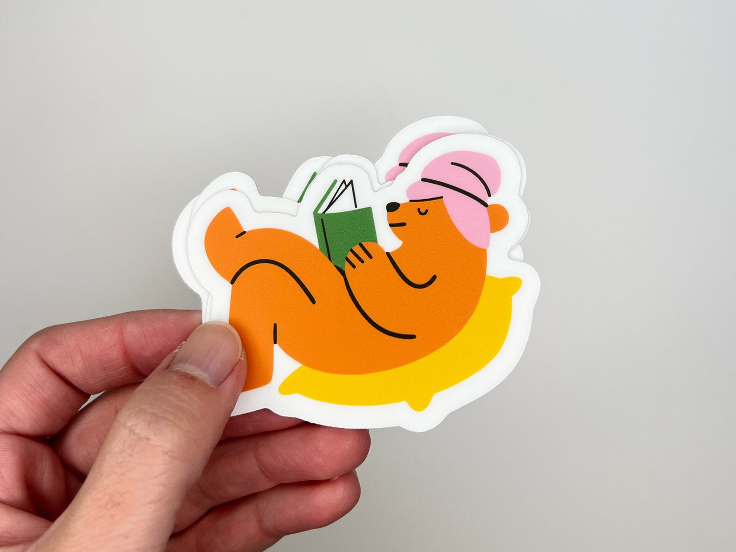 Hand holding a small stack of matte vinyl die cut stickers showing an orange bear wearing a pink towel wrapped around its head, lying on a yellow pillow while reading a green book. The cozy reading bear stickers are layered together against a light gray background.