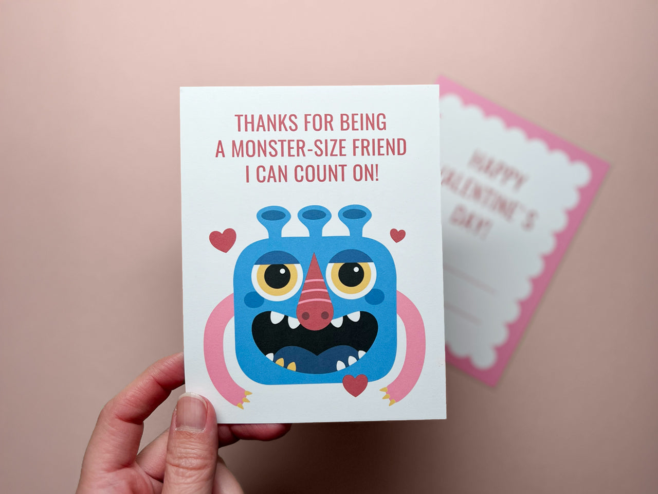 A hand holding a colorful printable card featuring a blue monster illustration with yellow eyes, red nose, and pink arms, surrounded by red hearts and red text. In the background shows the back of card slightly blurred.