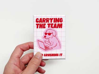 Hand holding a rectangular magnet with bold red text that says “Carrying the Team and Crushing It” featuring a pink illustration of a dog wearing sunglasses while working on a laptop over a light pink grid background.