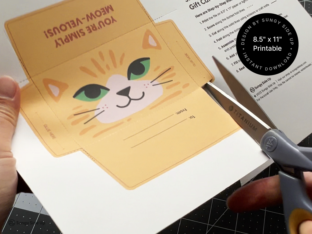 A person uses scissors to cut out the cat-themed gift card holder template printed on 8.5" x 11" paper. The sheet features a folded envelope design with an illustrated cat face, labeled tabs, and instructions. A circular label in the image confirms it's a printable instant download. This visual highlights the DIY nature of the product, showing that it’s easy to craft at home with basic tools and a standard printer.
