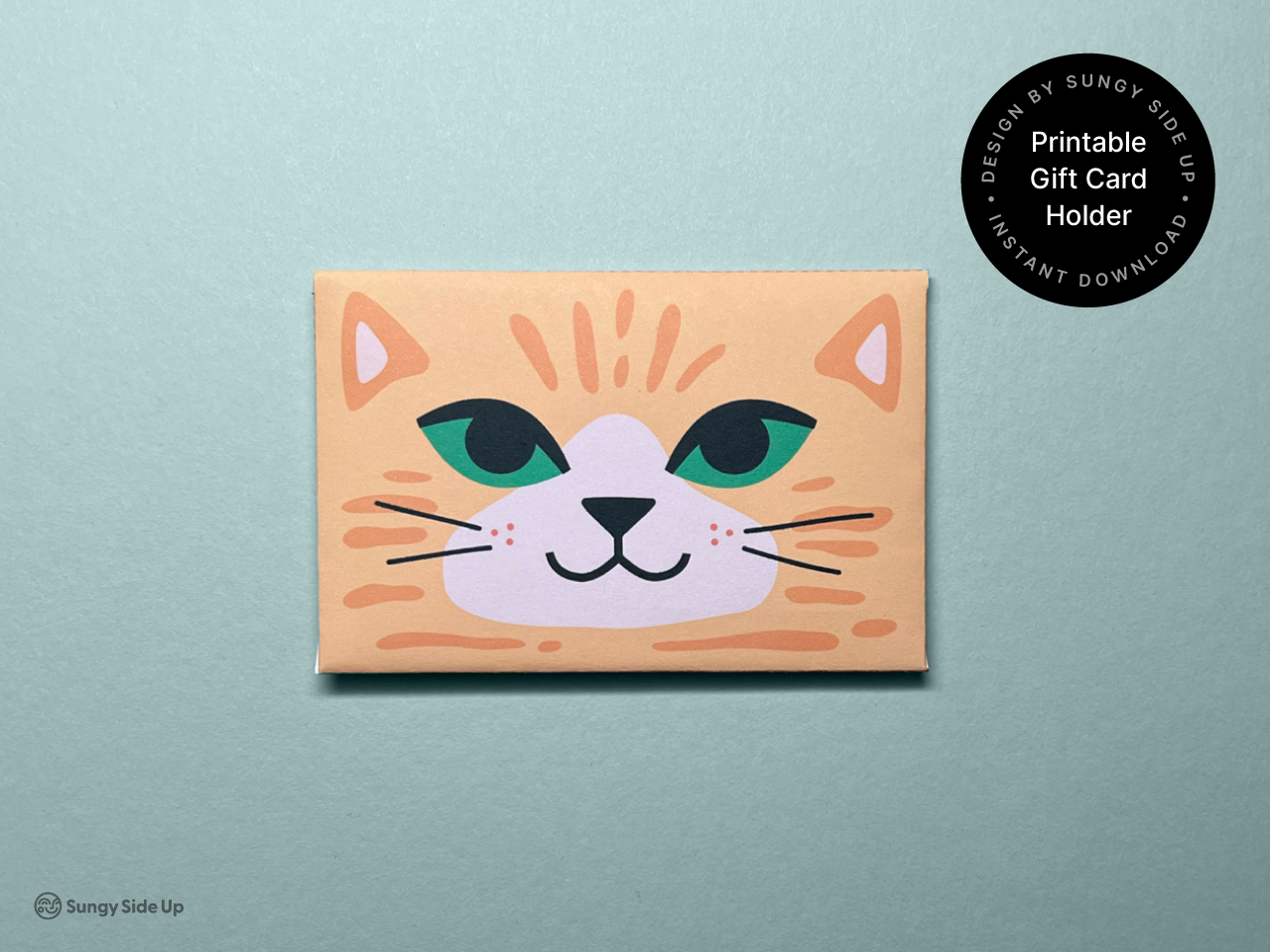 This close-up image showcases the front of the printable cat gift card holder. It features a charming orange cat face with green eyes and a slight smile. The folded envelope is set against a soft green background, with a round label that says “Printable Gift Card Holder • Instant Download.” This product is perfect for birthdays, thank-you cards, or any occasion that calls for a fun and personal handmade touch.