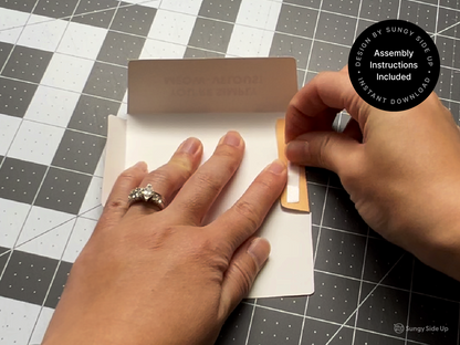 Two hands are shown folding and pressing down a tab on a printed DIY cat gift card holder. One hand holds the template steady while the other seals the flap with glue or tape. The envelope is partially assembled on a cutting mat, and a round badge notes that assembly instructions are included. This visual emphasizes the ease of at-home crafting with this instant download, perfect for gifting and classroom use.