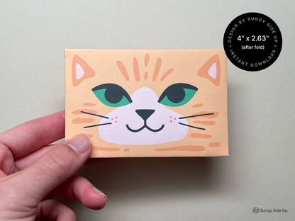 A close-up of a hand holding the completed folded envelope shows the front design of a cheerful orange cat with big green eyes and a soft smile. The envelope size is noted as 4" x 2.63" after folding, and a circular badge confirms it's an instant download. This DIY printable is designed for gifting with a handmade touch, offering a fun and personal way to present gift cards to kids, friends, or teachers.
