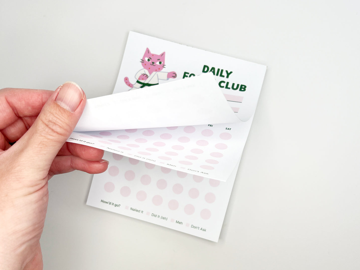Hand peeling back a sheet from the Daily Focus Club 4.25 x 5.5 monthly habit tracker notepad, revealing soft pink tracking dots and pink karate cat illustration underneath. Cute productivity notepad for goal setting, daily planning, and habit tracking with tear-off pages, designed by Sungy Side Up.