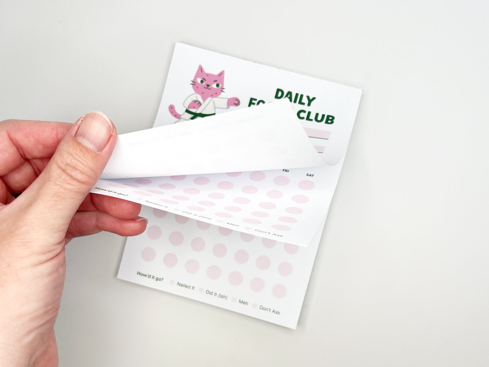 Hand peeling back a sheet from the Daily Focus Club 4.25 x 5.5 monthly habit tracker notepad, revealing soft pink tracking dots and pink karate cat illustration underneath. Cute productivity notepad for goal setting, daily planning, and habit tracking with tear-off pages, designed by Sungy Side Up.