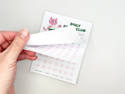 Hand peeling back a sheet from the Daily Focus Club 4.25 x 5.5 monthly habit tracker notepad, revealing soft pink tracking dots and pink karate cat illustration underneath. Cute productivity notepad for goal setting, daily planning, and habit tracking with tear-off pages, designed by Sungy Side Up.