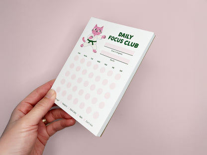 Hand holding Daily Focus Club monthly habit tracker notepad against soft pink background, featuring pink karate cat illustration, goal and month sections, and pastel daily tracking dots. Cute productivity stationery for goal setting, habit tracking, ADHD-friendly planning, and desk organization.