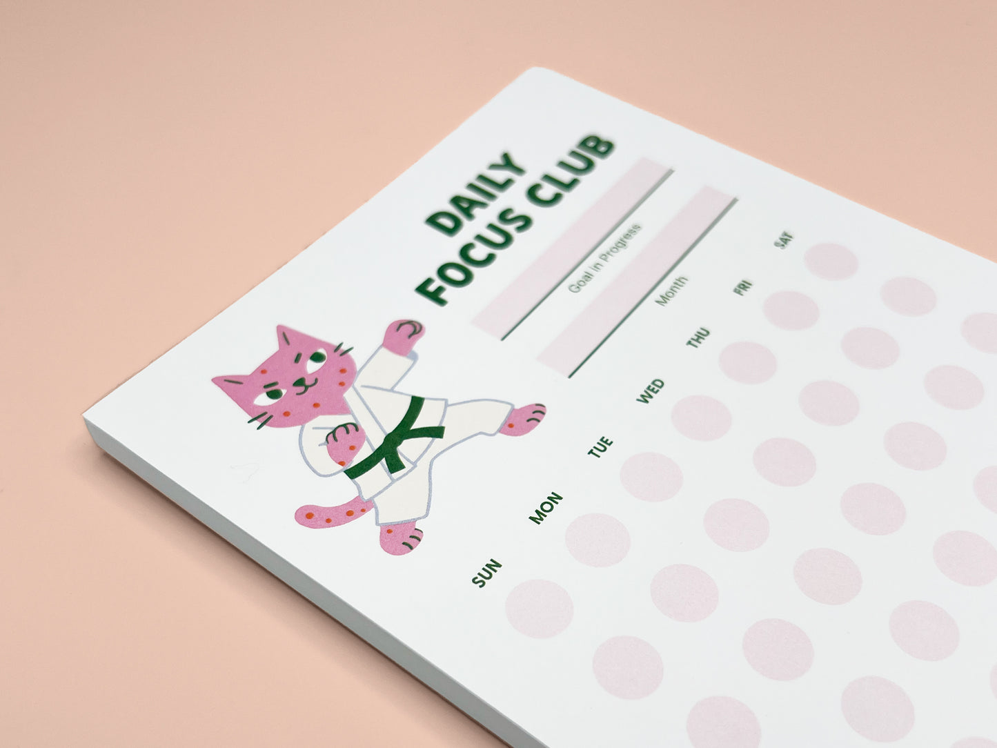 Close-up angled view of Daily Focus Club monthly habit tracker notepad showing pink karate cat illustration, goal-in-progress and month fields, and soft pink daily tracking dots labeled by day of week. Cute productivity stationery for goal setting, habit tracking, ADHD-friendly planning, and desk organization.