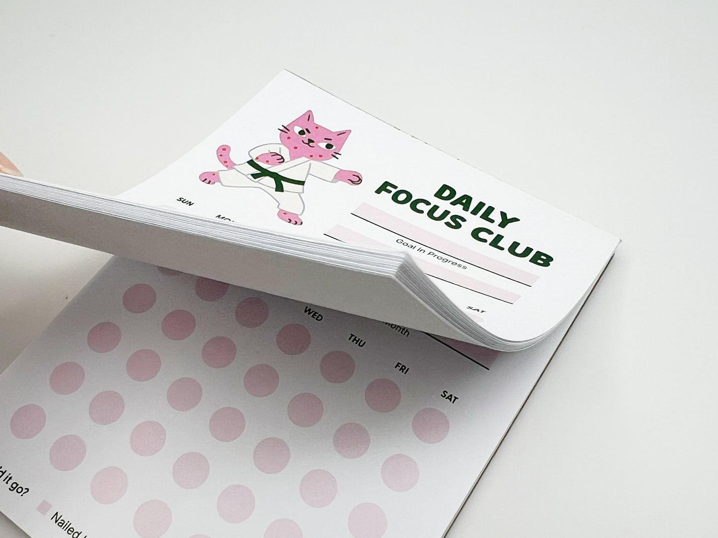 Stacked tear-off pages of Daily Focus Club monthly habit tracker notepad showing pink karate cat illustration, goal-in-progress section, and soft pink daily tracking dots labeled by day of week. Cute productivity stationery for goal setting, habit tracking, ADHD-friendly planning, and desk organization.