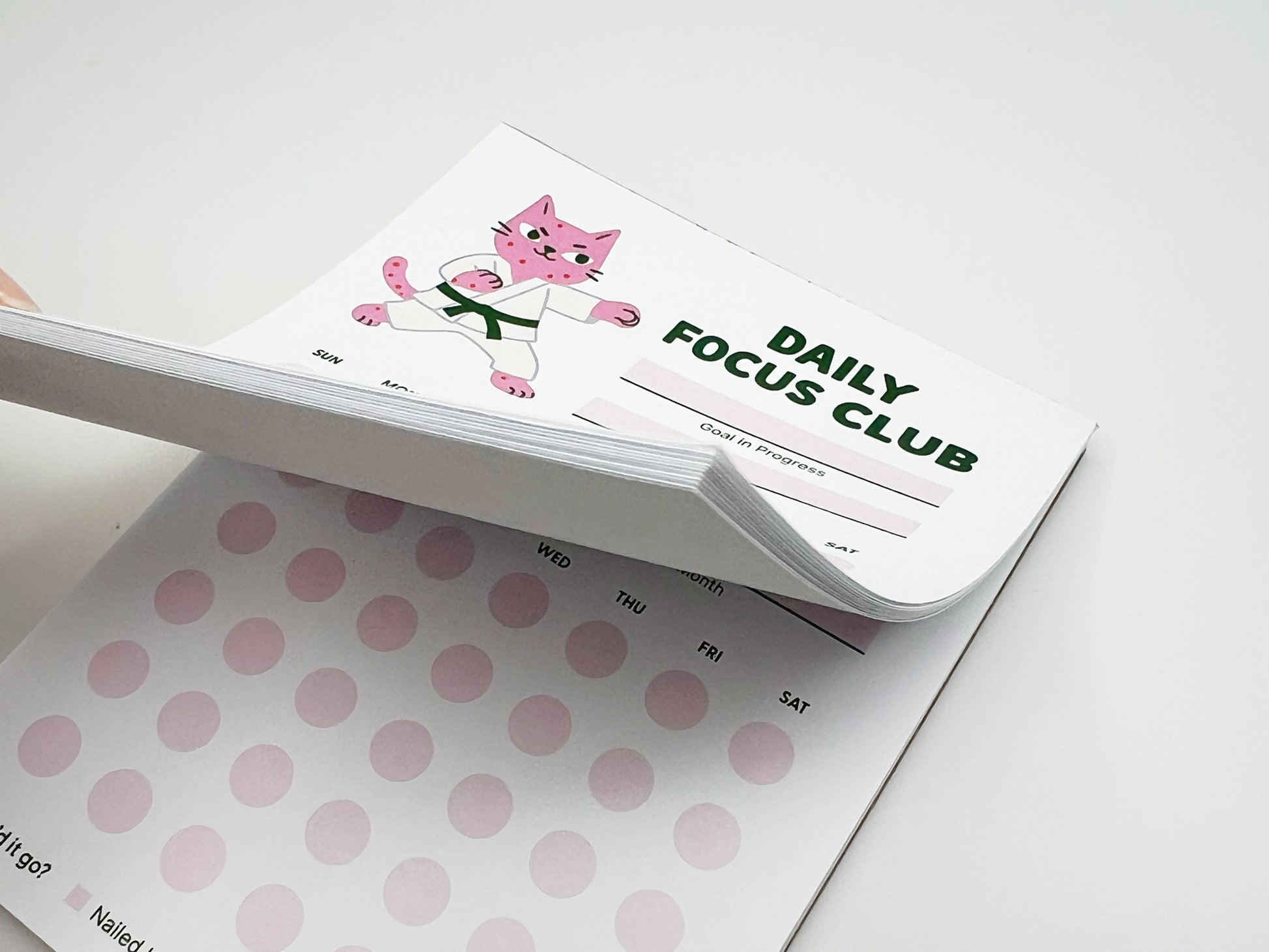 Stacked tear-off pages of Daily Focus Club monthly habit tracker notepad showing pink karate cat illustration, goal-in-progress section, and soft pink daily tracking dots labeled by day of week. Cute productivity stationery for goal setting, habit tracking, ADHD-friendly planning, and desk organization.