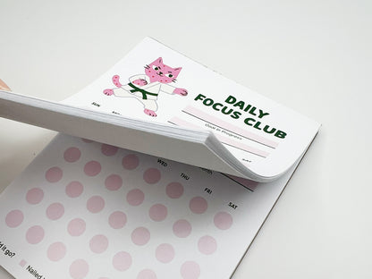 Stacked tear-off pages of Daily Focus Club monthly habit tracker notepad showing pink karate cat illustration, goal-in-progress section, and soft pink daily tracking dots labeled by day of week. Cute productivity stationery for goal setting, habit tracking, ADHD-friendly planning, and desk organization.